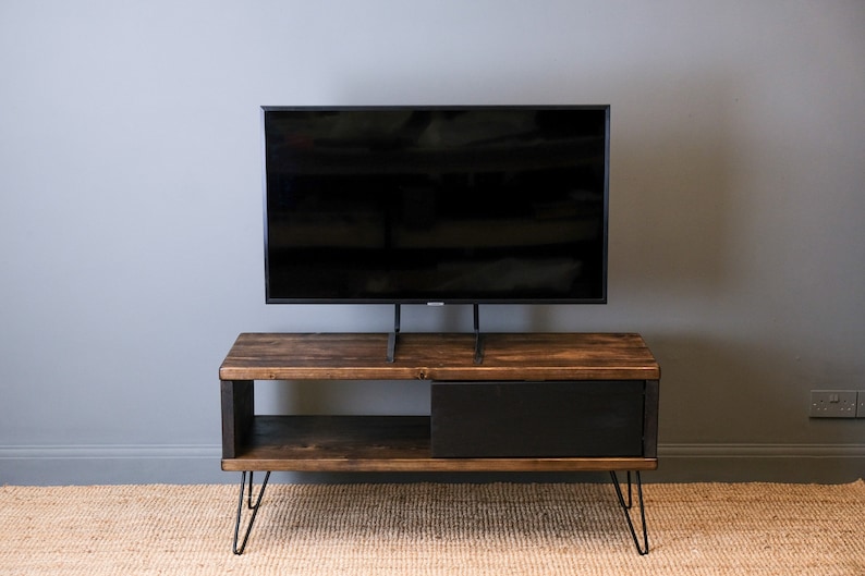 May include: A dark brown wooden TV stand with a black drawer and a shelf. The stand has hairpin legs and is supporting a black TV on a stand.