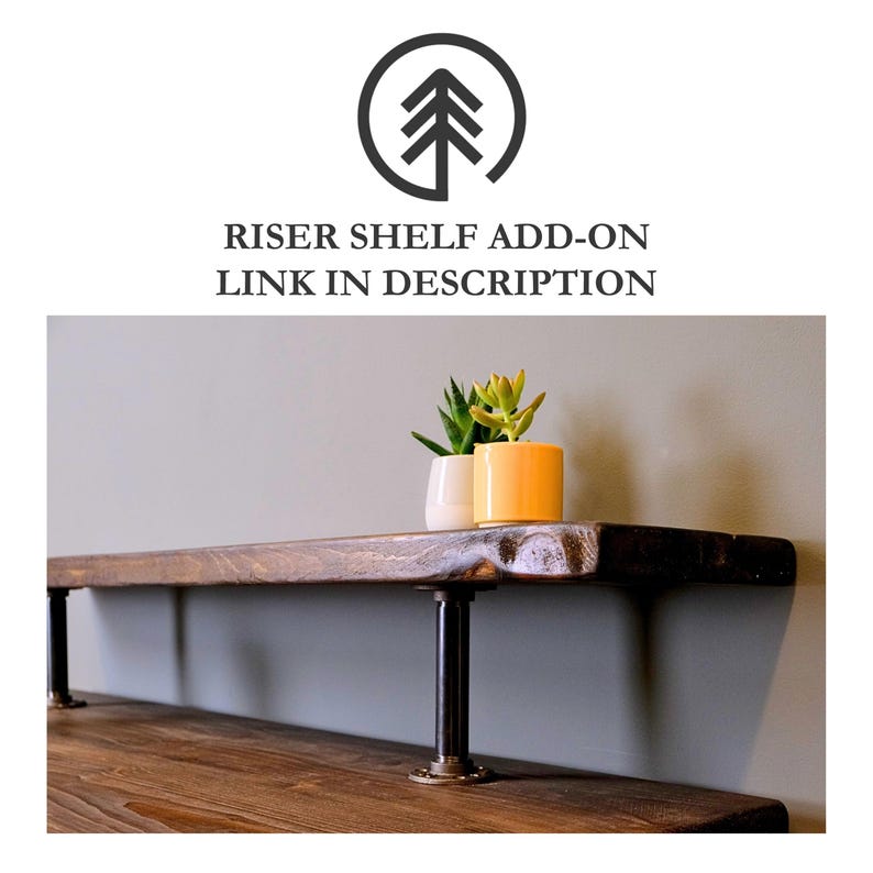 A wooden monitor stand with black metal pipe supports. The shelf is mounted to a desk and has two small potted plants on top.