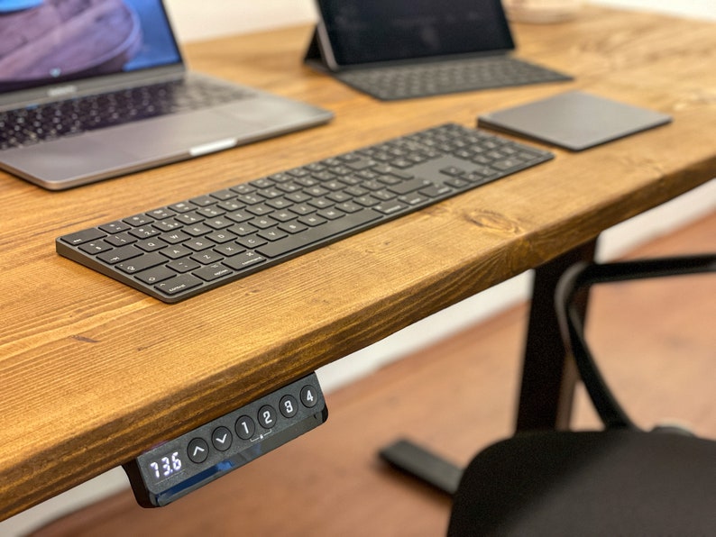 A wooden desk with a black keyboard, a black trackpad, and a black laptop. The desk has a black control panel with a digital display that reads "73.6". The control panel has buttons numbered 1 to 4 and up and down arrows.