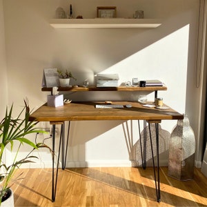 Bright and serene home workspace featuring a dark wood desk with black hairpin legs and a raised shelf. The desk is styled with a calendar, tissue box, wireless keyboard, candles, and a small plant, with sunlight casting soft shadows across the wooden floor and wall. A sculptural lamp and leafy plant add warmth and character to the cosy corner.