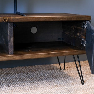 May include: A dark brown wooden TV stand with a cabinet door open, revealing a shelf inside. The stand has black hairpin legs and is sitting on a rug.
