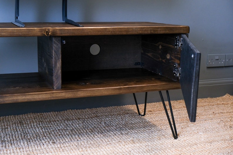 May include: A dark brown wooden TV stand with a cabinet door open, revealing a shelf inside. The stand has black hairpin legs and is sitting on a rug.