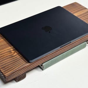 May include: A dark-colored laptop rests on a stained wooden laptop stand. The stand is rectangular with a raised design and small feet. A green book is placed between the stand and the laptop. The laptop has a black Apple logo.