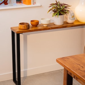 Slim wooden console table with black steel square legs, placed over a radiator and styled with ceramic bowls, a plant, and a lamp in a warm, modern interior.