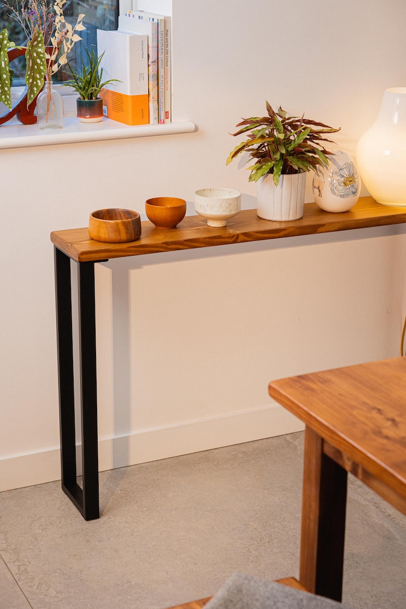 Slim wooden console table with black steel square legs, placed over a radiator and styled with ceramic bowls, a plant, and a lamp in a warm, modern interior.