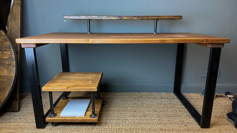 Puede incluir: Un escritorio de madera con patas de metal negro y un estante debajo. El escritorio tiene una tapa de madera y una barra de metal en la parte superior.