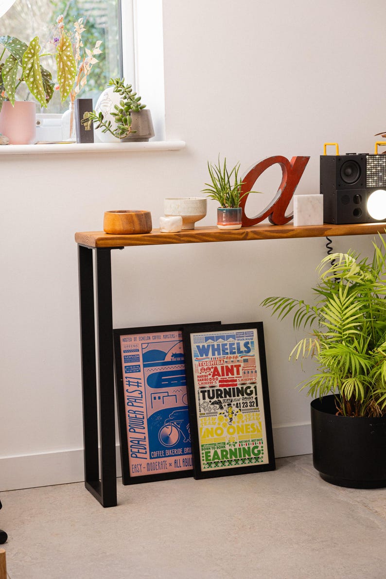 Wooden console table with black square metal legs, styled as a radiator shelf in a modern hallway with plants, books, and decorative objects on top.