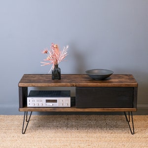 May include: A dark wood TV stand with a black drawer and a shelf. The stand has hairpin legs and is sitting on a woven rug. A small vase with dried flowers and a black bowl are on the top of the stand.