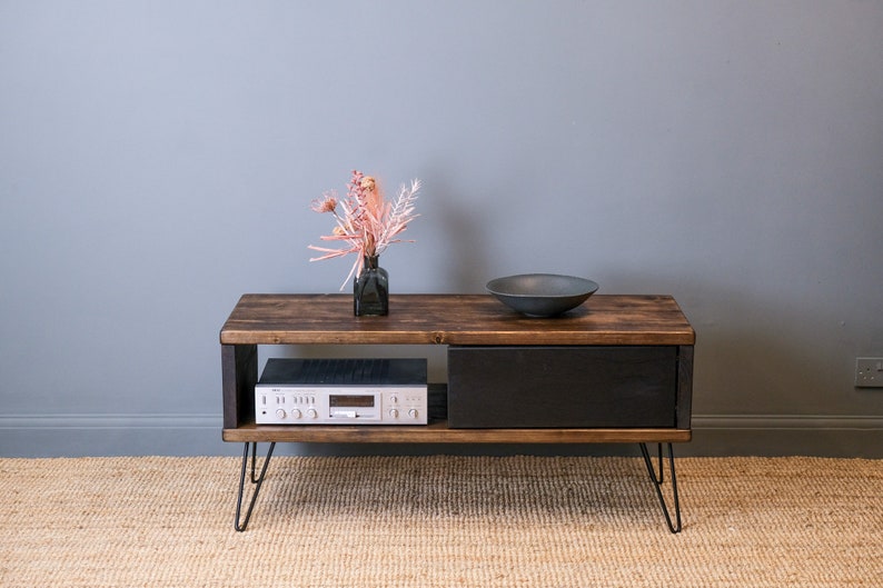 May include: A dark wood TV stand with a black drawer and a shelf. The stand has hairpin legs and is sitting on a woven rug. A small vase with dried flowers and a black bowl are on the top of the stand.