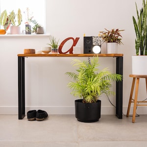 Wooden console table with black square metal legs, styled as a radiator shelf in a modern hallway with plants, books, and decorative objects on top.