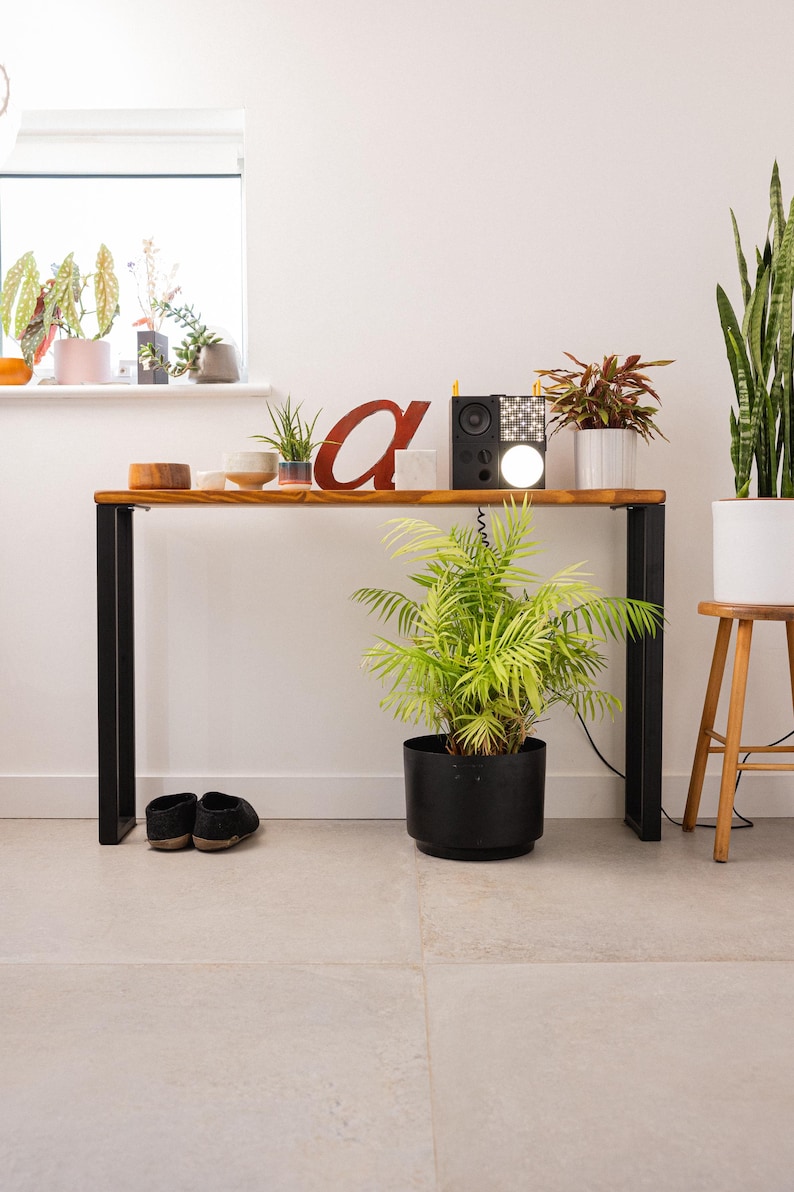 Wooden console table with black square metal legs, styled as a radiator shelf in a modern hallway with plants, books, and decorative objects on top.