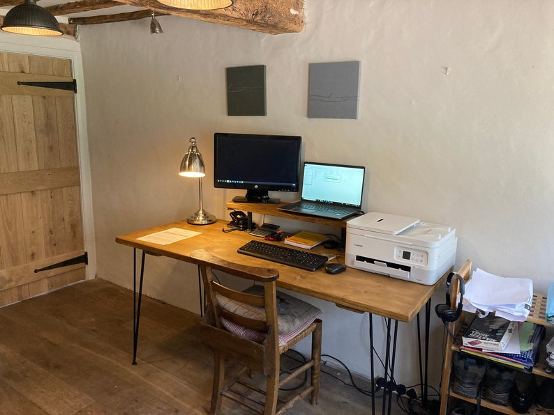 Rustic home office with a natural wood desk and black hairpin legs, paired with a vintage wooden chair and a woven cushion. The workspace features a desktop monitor, laptop, printer, and reading lamp, all set against a textured white wall with exposed ceiling beams. A functional and characterful setup for a cosy work-from-home routine.