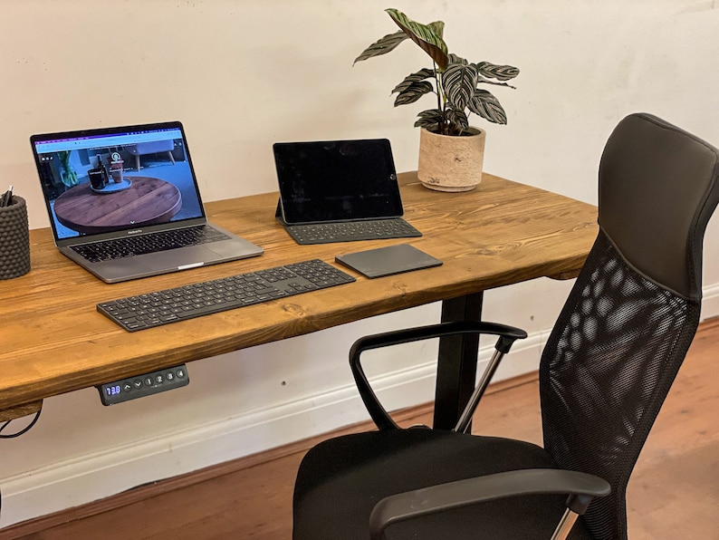 A wooden standing desk with a black mesh office chair. The desk has a laptop, keyboard, tablet, and a plant on it. The desk has a control panel for adjusting the height.