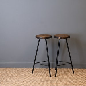 Two solid wood bar stools with black metal legs on a woven rug against a grey wall.