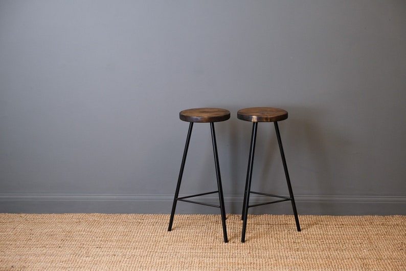 Two solid wood bar stools with black metal legs on a woven rug against a grey wall.