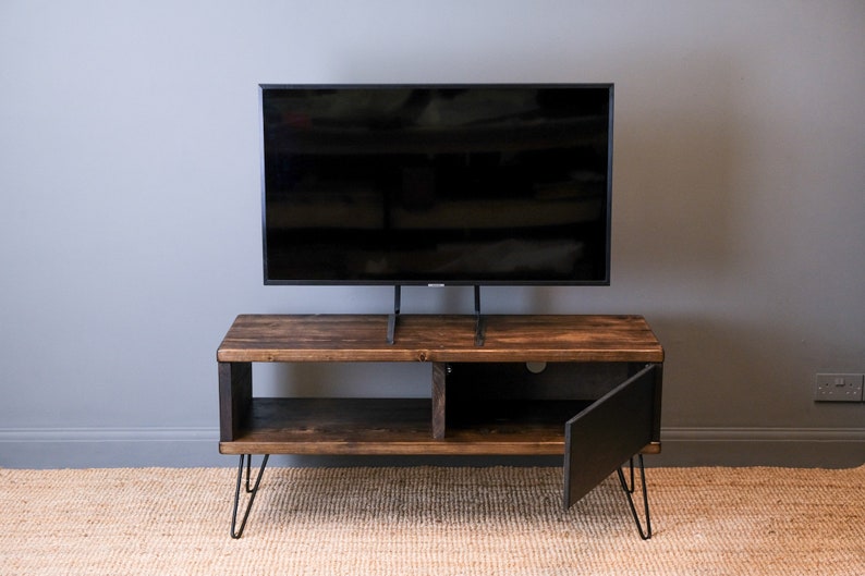 May include: A dark wood TV stand with a black door and hairpin legs. The stand has a shelf and a space for a TV. The TV is on a black stand.