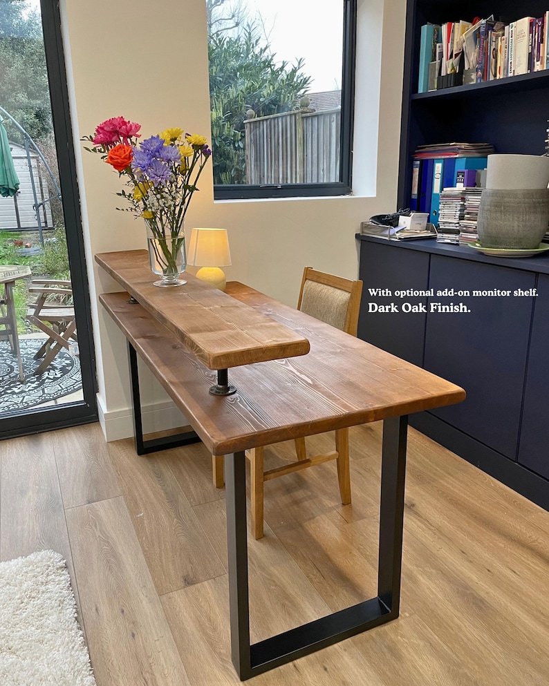 Solid wooden desk with a medium to dark finish and black steel legs. It features an optional monitor shelf add-on and it's styled with a vase of flowers.