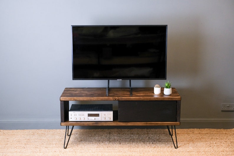 May include: A dark brown wooden TV stand with a black drawer and a black metal frame. The stand has hairpin legs and is sitting on a light brown rug. A flat screen TV is sitting on top of the stand.