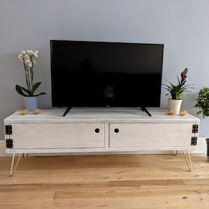 May include: White wooden TV stand with two doors and gold hairpin legs. The stand has a distressed white finish and is sitting on a wood floor.