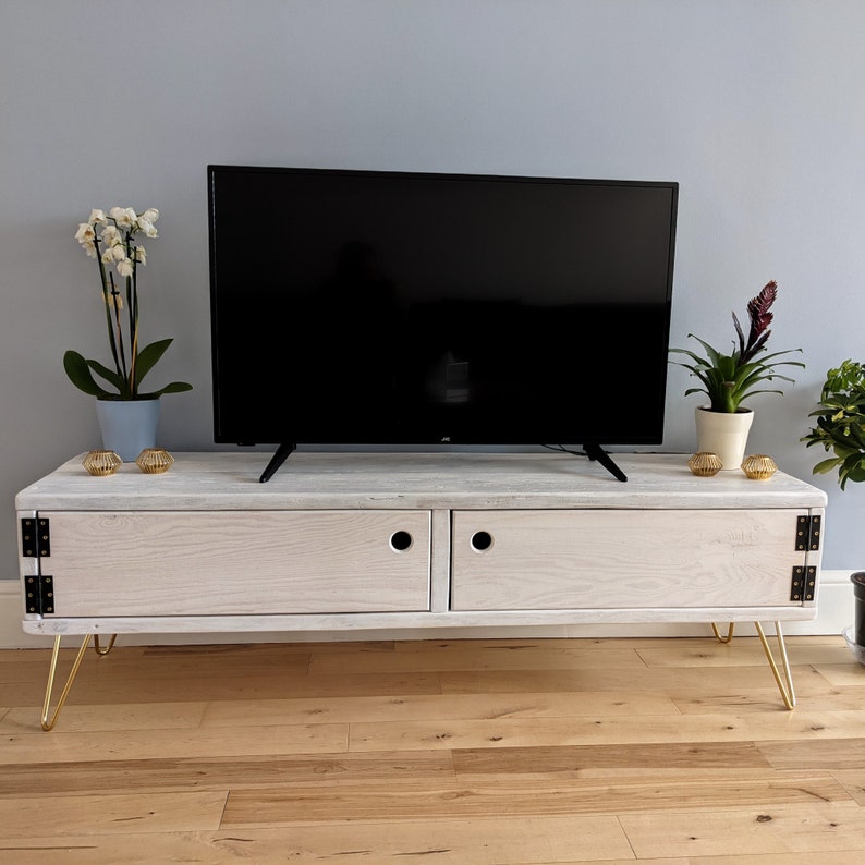 May include: White wooden TV stand with two doors and gold hairpin legs. The stand has a distressed white finish and is sitting on a wood floor.