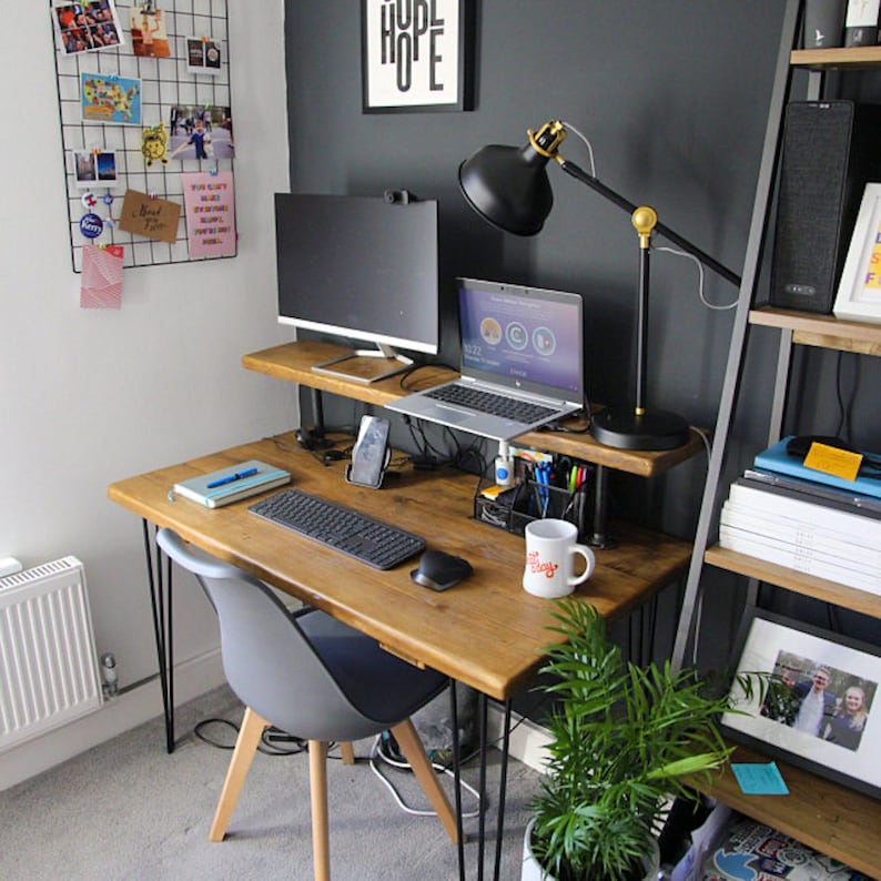 Compact home office desk with a natural wood top and black hairpin legs, featuring a raised shelf holding two monitors and a desk lamp. The workspace includes a grey chair with wooden legs, a keyboard, mouse, notebooks, and various office accessories. A wire memo board with photos is mounted on the white wall, and a shelving unit with books and decor stands on the right. A small green plant sits on the floor in the foreground.