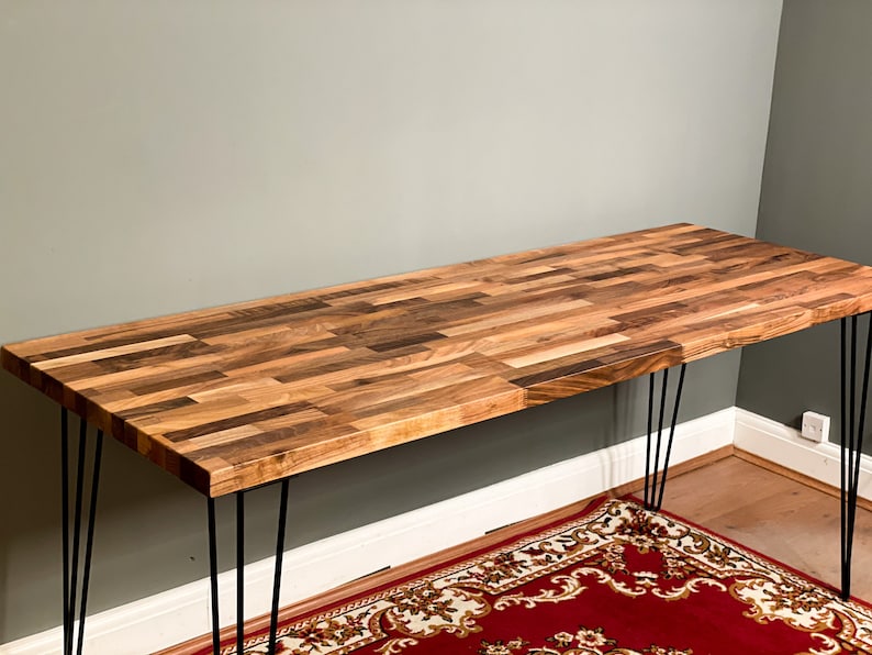 May include: A wooden desk with a natural wood finish and black hairpin legs. The desk is made of multiple pieces of wood that are arranged in a parquet pattern.
