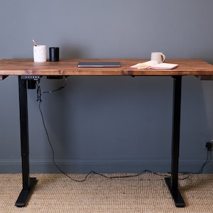 A wooden standing desk with black metal legs and a power outlet on the underside. The desk has a laptop, a cup, and a notebook on top.