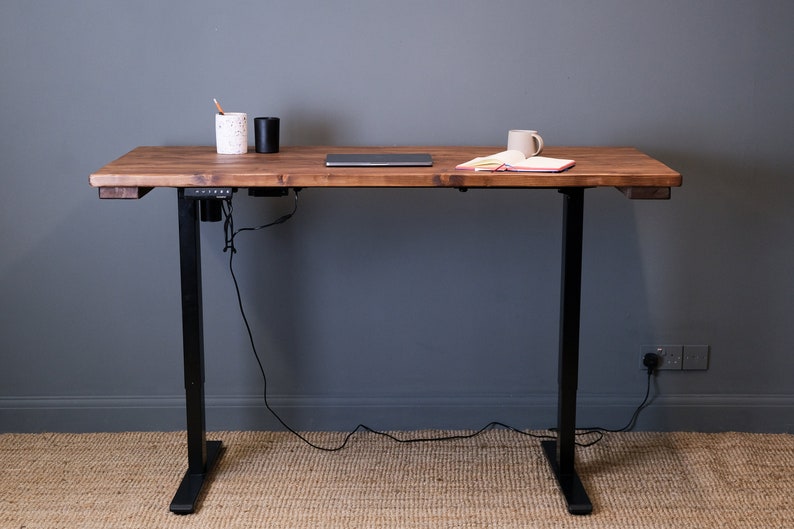 A wooden standing desk with black metal legs and a power outlet on the underside. The desk has a laptop, a cup, and a notebook on top.