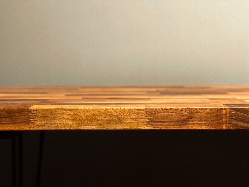 May include: A close-up of a wooden table top with a light brown finish. The table top is made of multiple pieces of wood that are arranged in a checkered pattern. The table top is in focus, while the background is blurred.