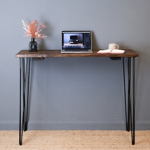 May include: A dark brown wooden desk with black hairpin legs. A laptop computer, a notebook, and a mug are on the desk. A vase with dried flowers is on the left side of the desk.