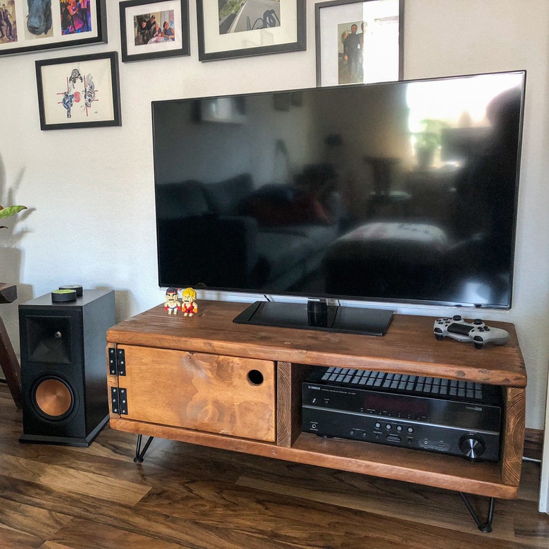 May include: A rustic wooden TV stand with a dark brown finish and black metal legs. The stand has a cabinet door with a cut-out handle and an open shelf for electronics. A flat screen TV is mounted on top of the stand, and a stereo receiver is on the shelf.