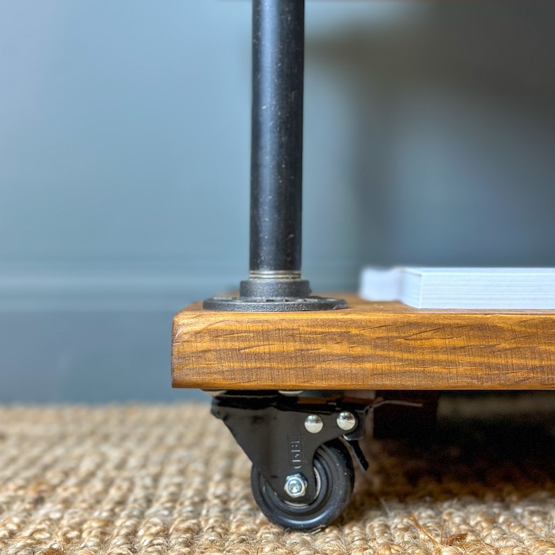 Puede incluir: Una estanter&iacute;a de madera con patas de tubo de metal negro y ruedas negras. La estanter&iacute;a est&aacute; te&ntilde;ida de un color marr&oacute;n oscuro y tiene una pila de papel blanco en la parte superior.