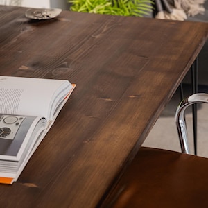 Close-up of a dark-stained wooden desk showing its smooth grain and natural knots, with a chrome-framed brown leather chair tucked underneath. An open design book and a small ceramic dish rest on the surface, while soft furnishings and greenery in the background add a relaxed, homely feel.