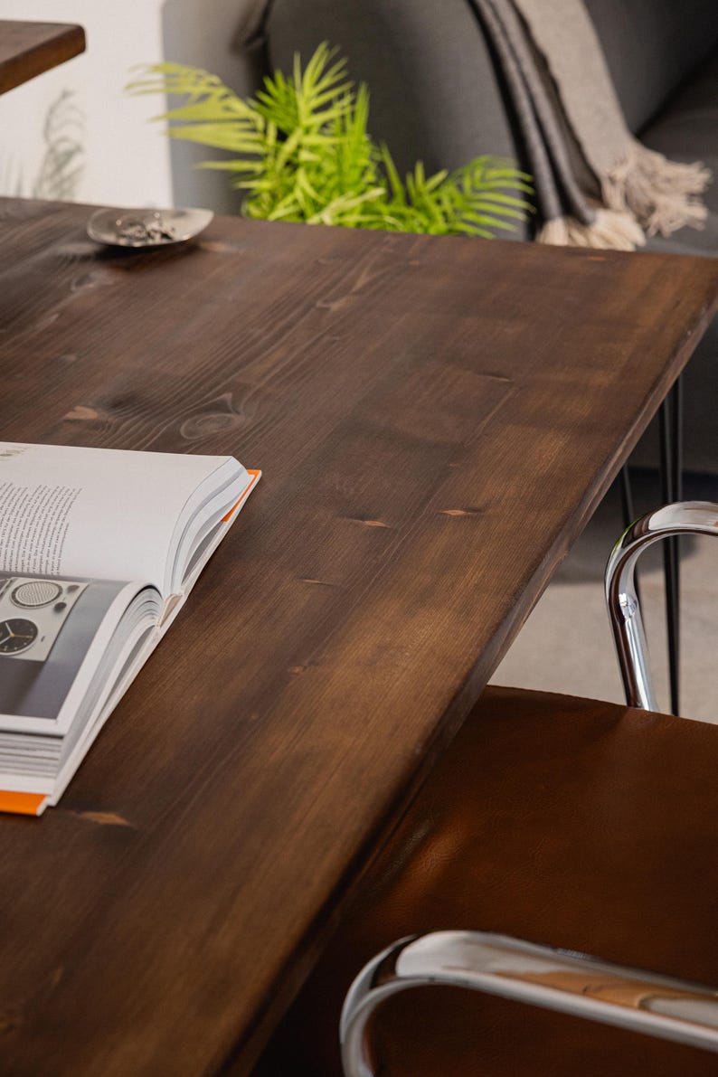 Close-up of a dark-stained wooden desk showing its smooth grain and natural knots, with a chrome-framed brown leather chair tucked underneath. An open design book and a small ceramic dish rest on the surface, while soft furnishings and greenery in the background add a relaxed, homely feel.