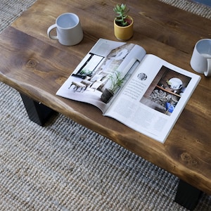 May include: A rustic wooden coffee table with a live edge design. The table has a natural wood finish and is decorated with two white mugs, a magazine, and a small potted succulent plant. The table is sitting on a woven rug.