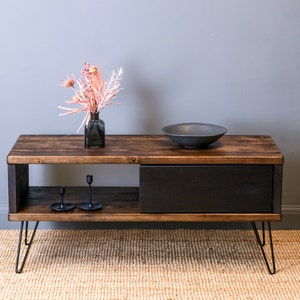 May include: A dark wood coffee table with a black drawer and open shelf. The table has hairpin legs and is decorated with a vase of pink flowers and a black bowl.