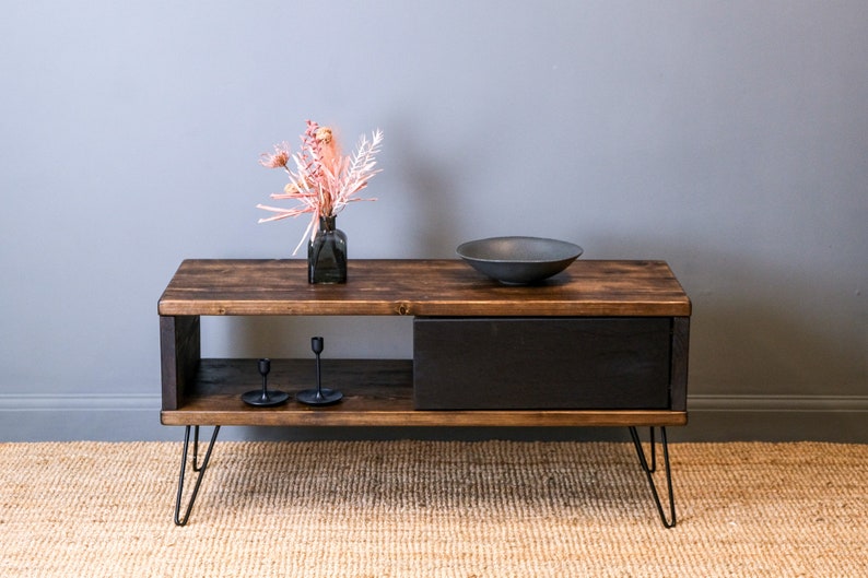 May include: A dark wood coffee table with a black drawer and open shelf. The table has hairpin legs and is decorated with a vase of pink flowers and a black bowl.