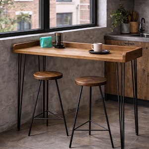 Solid wood kitchen breakfast bar table with black hairpin legs and two stools beside a large window in a modern kitchen.