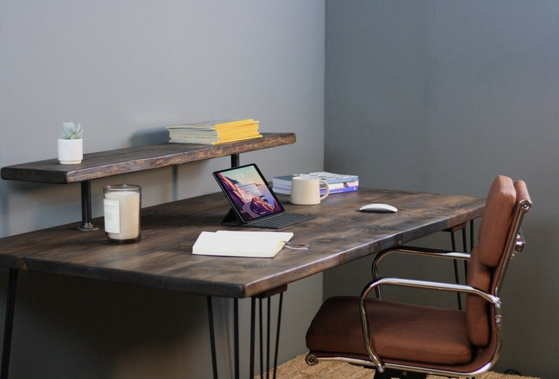 Cosy workspace with a dark wood desk and black hairpin legs, styled with a tablet, books, candle, notebook, and a ceramic mug. A raised shelf holds magazines and a small potted plant, while a brown leather swivel chair adds comfort. Set against a soft grey wall with a relaxed, creative vibe.