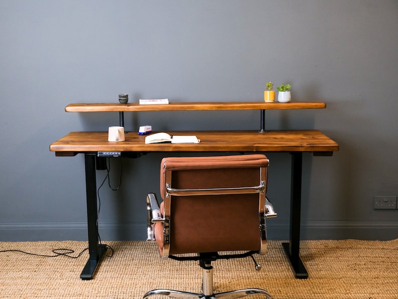 A wooden sit stand desk with a black metal frame and a brown leather office chair. The desk has a top shelf and a lower shelf. There are two small potted plants on the top shelf.