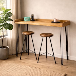 Solid wood breakfast bar table with black hairpin legs and two stools by a window, styled with coffee and a plant in a bright living room space.