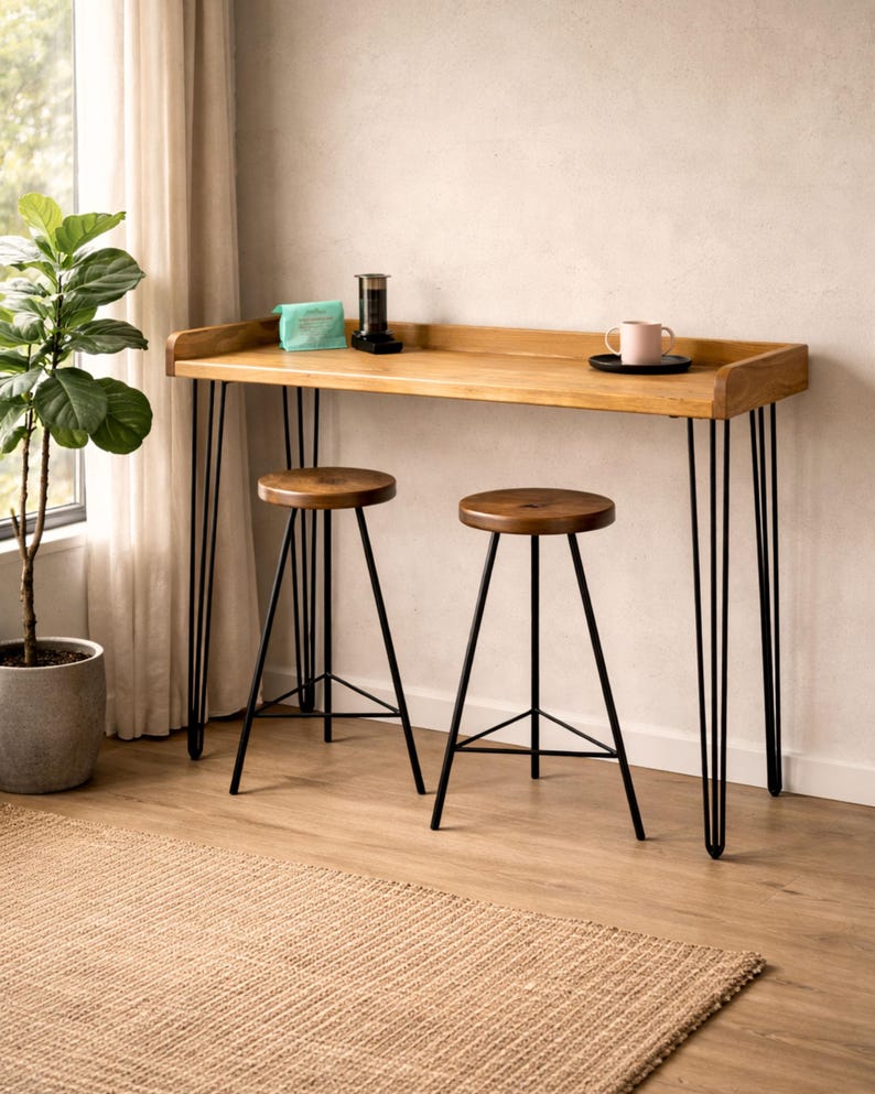 Solid wood breakfast bar table with black hairpin legs and two stools by a window, styled with coffee and a plant in a bright living room space.