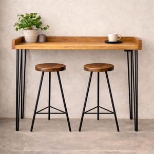 Solid wood breakfast bar table with black hairpin legs and two stools, styled with a plant and coffee cup against a neutral wall.