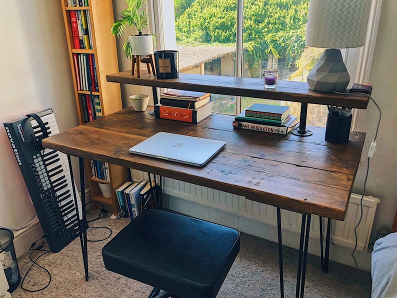 Rustic wooden desk with a shelf, featuring a weathered dark wood surface and black hairpin legs, placed by a window. On the desk are a closed laptop, books, a candle, and a table lamp with a geometric base. The shelf holds a small plant, candle, and more books. A keyboard stands vertically against the wall, and a tall bookshelf filled with colourful books is visible on the left. Natural light streams in from the window behind.