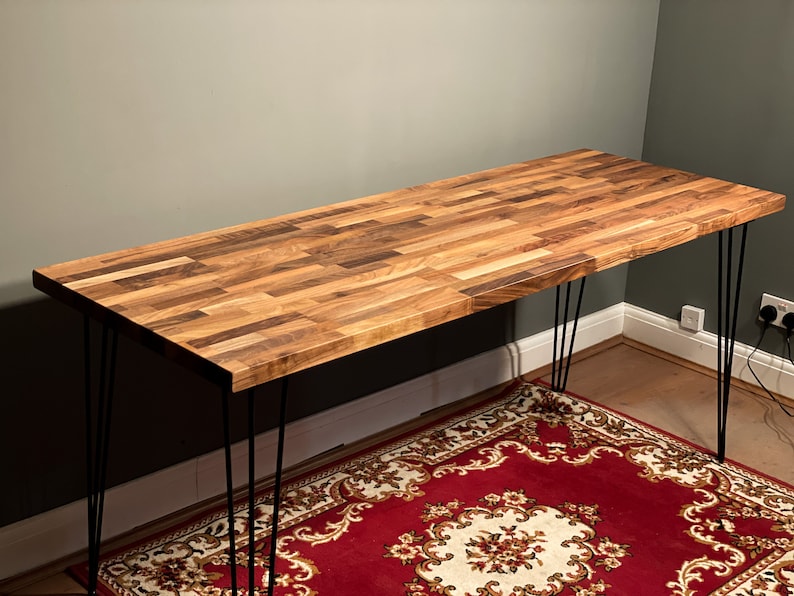 May include: A wooden desk with a natural wood finish and black hairpin legs. The desk is sitting on a red and white patterned rug.