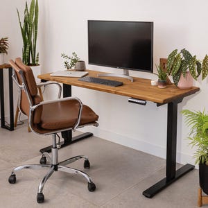 May include: A brown leather office chair with chrome legs is positioned in front of a wooden desk with a black base. A computer monitor, keyboard, and laptop are on the desk. Potted plants add greenery to the home office.