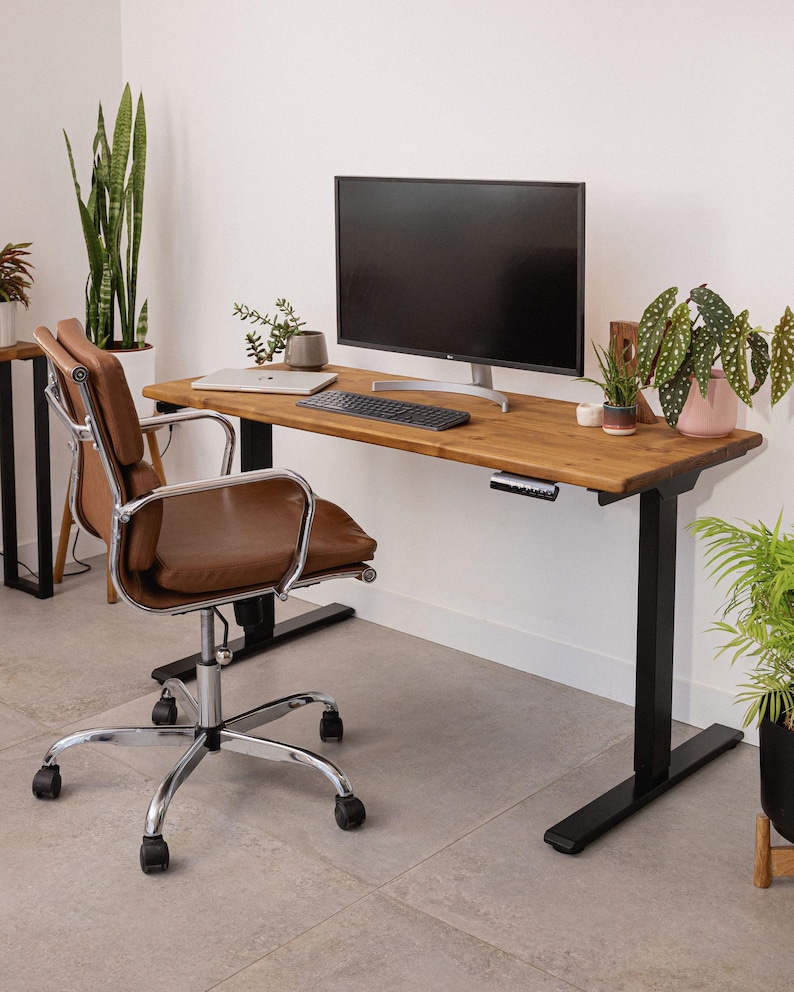 Height adjustable solid wood desk with black metal legs, set against a white wall with monitor, keyboard, laptop and indoor plants, paired with a brown leather office chair.