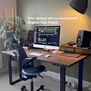Solid wooden desk with dark finish and black steel legs. Featuring a monitor shelf. In a dark green wall room with plants and low ambient light.