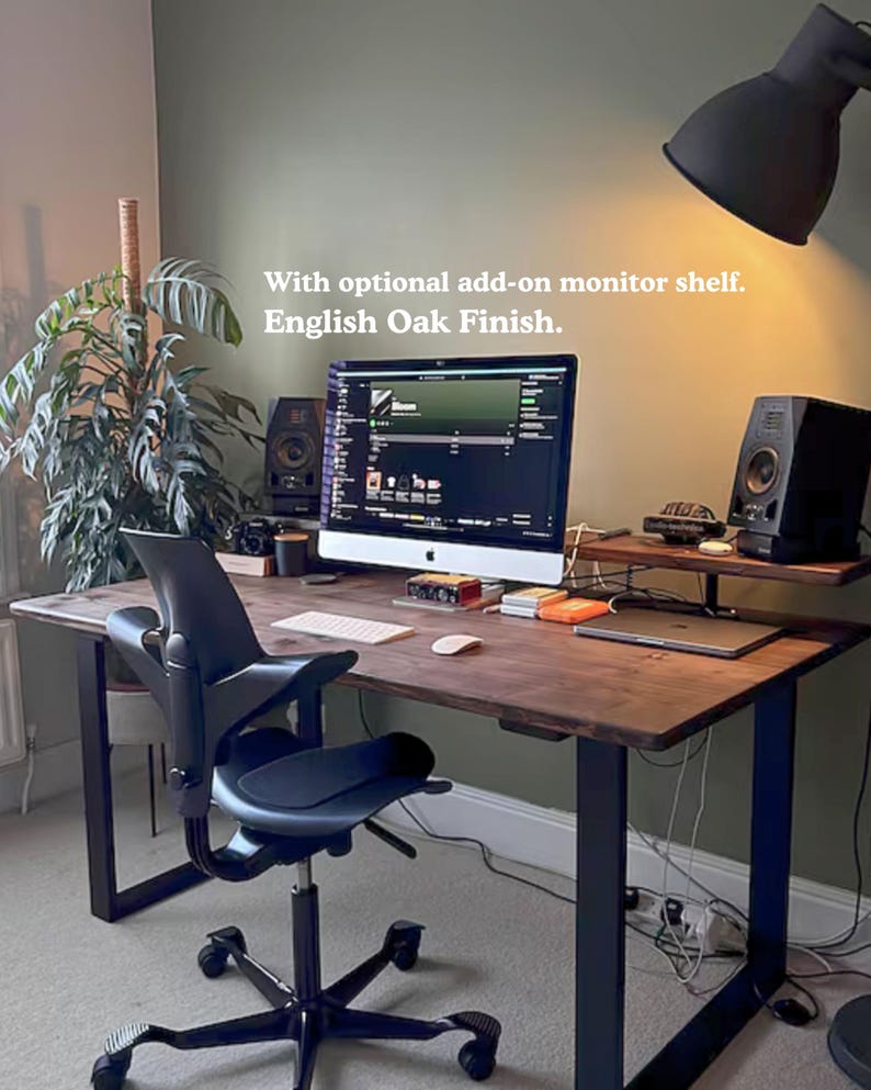 Solid wooden desk with dark finish and black steel legs. Featuring a monitor shelf. In a dark green wall room with plants and low ambient light.