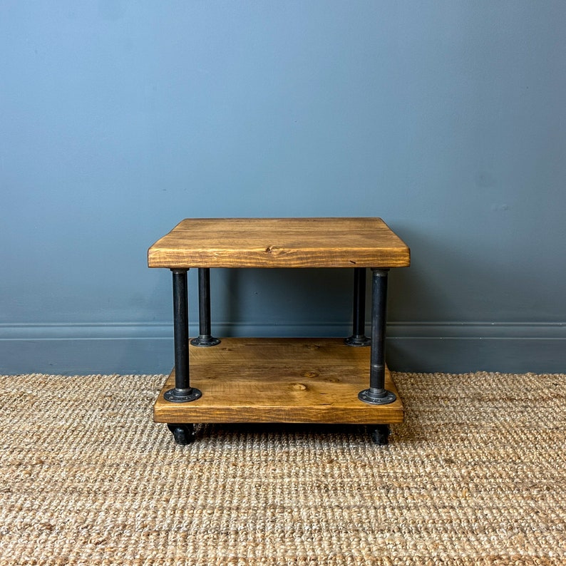 Puede incluir: Una mesa de caf&eacute; de madera r&uacute;stica con un marco de metal negro y dos estantes. La mesa tiene un acabado de madera desgastada y est&aacute; soportada por cuatro patas de metal negro.